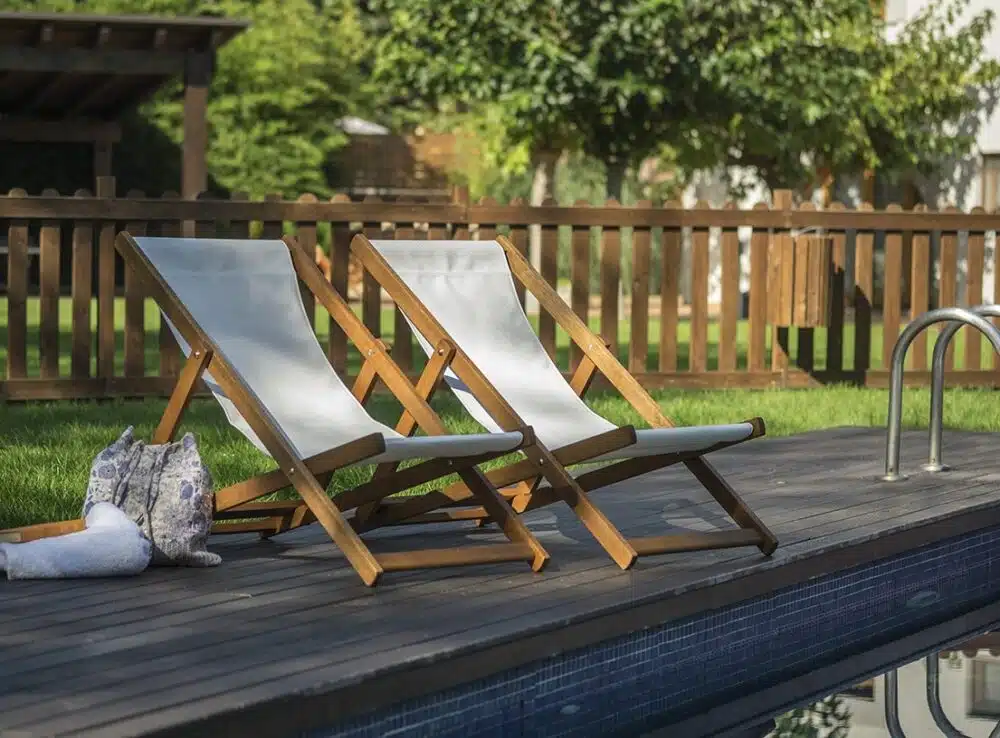 Se observan dos mini tumbonas con estructura de madera y tejido de color blanco, las mini tumbonas se ven en un jardin junto a una piscina