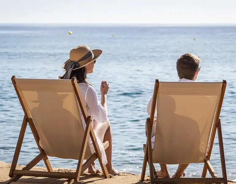 Se puede observar en la imagen dos mini tumbonas en color natural en la playa con dos personas sentadas encima