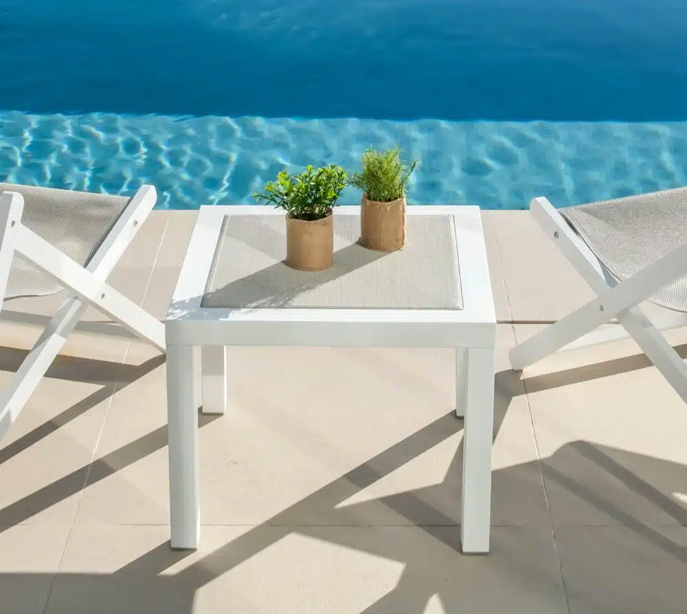 Mesa blanca con el centro del tablero hecho de tejido color natural, la mesa esta junto a dos minitumbonas en una piscina