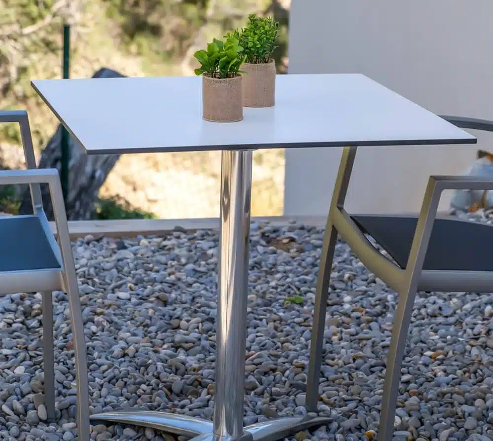 Imagen de una mesa moderna con superficie cuadrada de color gris claro y base central de metal cromado con cuatro patas inclinadas. La mesa tiene un diseño minimalista y contemporáneo, ideal para bares o restaurantes modernos