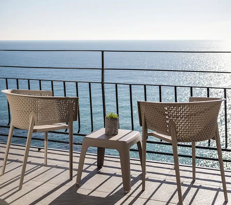 Imagen de un balcón con vista al mar mostrando dos sillas de mimbre modernas y una mesa pequeña con una planta en maceta, sobre un piso de baldosas, con una barandilla metálica y el horizonte marino en un día soleado