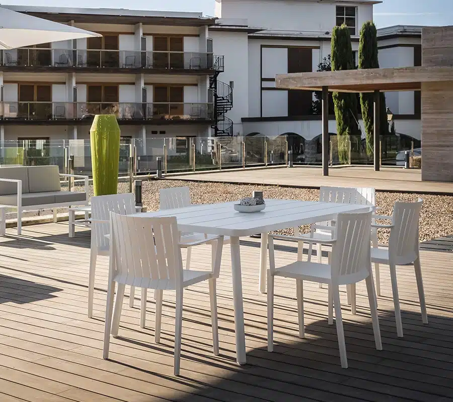 Silla de exterior blanca con reposabrazos - Modelo Vera. Diseño minimalista y contemporáneo, perfecto para ambientes al aire libre como terrazas y patios. La silla se encuentra en un entorno rodeado de vegetación exuberante, con un suelo de madera natural