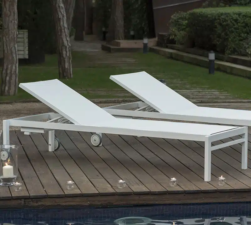 En la imagen se muestran dos tumbonas de diseño elegante con chasis de aluminio y con tejido de color blanco con ruedas para poder desplazarlas, se ven junto a una piscina y con el cesped de fondo