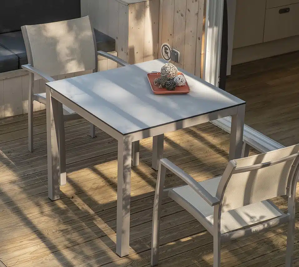 La imagen muestra un área de comedor sencilla y moderna, con una mesa blanca y dos sillas. Las sillas tienen un diseño de marco metálico elegante con asientos y respaldos blancos. Sobre la mesa, hay un pequeño objeto decorativo que parece ser una planta o arreglo floral en un cuenco naranja. La iluminación proviene de la luz natural del sol, creando sombras suaves en el suelo de madera, lo que sugiere que la habitación tiene ventanas grandes o puertas de vidrio cercanas