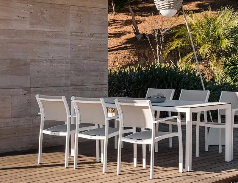 La imagen muestra un conjunto de muebles de exterior compuesto por sillas blancas y una mesa blanca. Están ubicados en una terraza con piso de madera. El fondo revela un entorno natural con plantas y árboles, así como una pared de concreto a la izquierda. La iluminación sugiere que es al aire libre durante el día