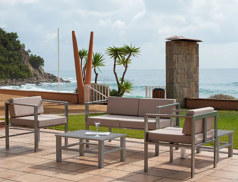 La imagen muestra un ambiente de patio al aire libre con vista a una playa. Hay un conjunto de muebles de patio modernos, incluyendo un sofá, dos sillones y una mesa de café con un vaso encima. En el fondo, se puede ver el océano