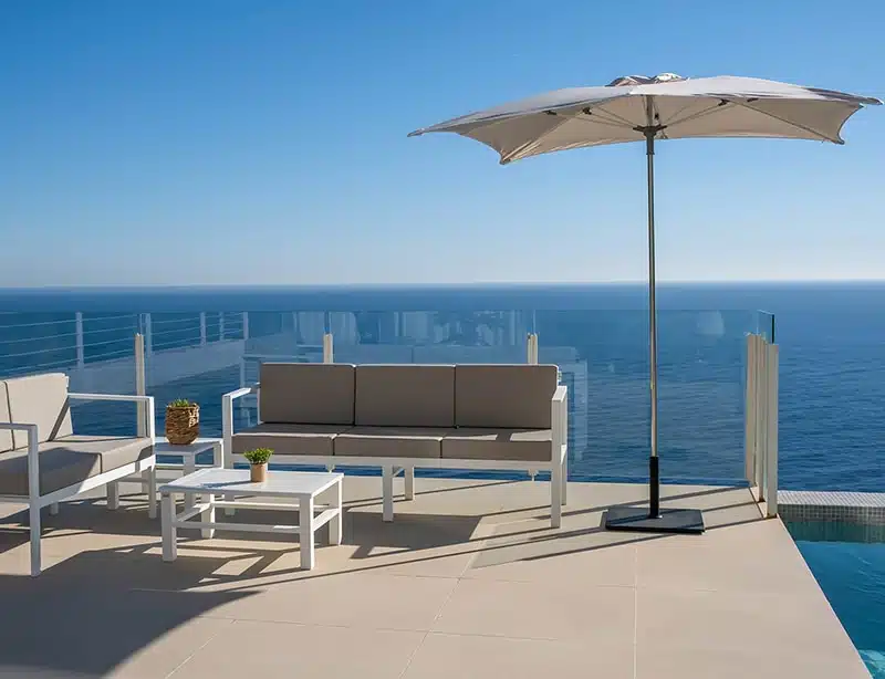 Terraza moderna con vista al mar, equipada con un sofá de 3 plazas color beige, otro sofa pero de 2 plazas, una mesa de centro blanca y un parasol grande abierto. La barandilla de cristal proporciona una vista despejada del horizonte marino bajo un cielo despejado