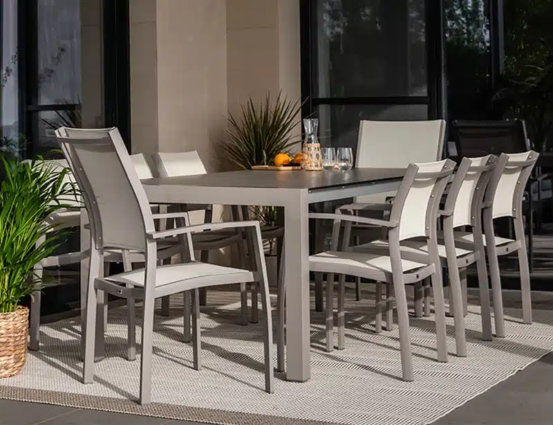 Conjunto de comedor al aire libre con mesa rectangular gris oscuro y ocho sillas a juego en terraza moderna, suelo de baldosas claras, alfombra tejida en tonos neutros y plantas verdes de fondo