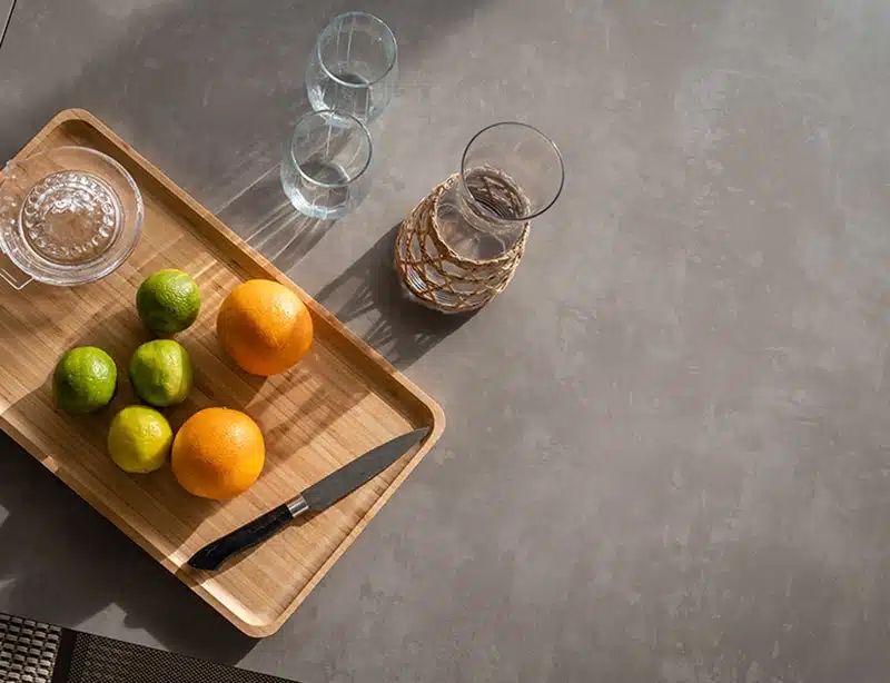 Bandeja de madera con dos naranjas, tres limas y un cuenco pequeño con cubos de azúcar sobre una superficie gris texturizada, junto a un vaso de agua decorado con patrón geométrico dorado y dos vasos boca abajo