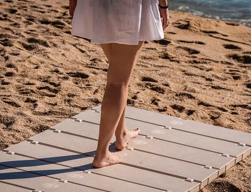 La mitad inferior de una persona de pie en una pasarela de playa en la arena de una playa. La persona lleva una prenda blanca que llega justo por encima de las rodillas. Un pie está colocado en la pasarela mientras el otro pie está ligeramente levantado, tocando solo con los dedos de los pies, dando la impresión de movimiento