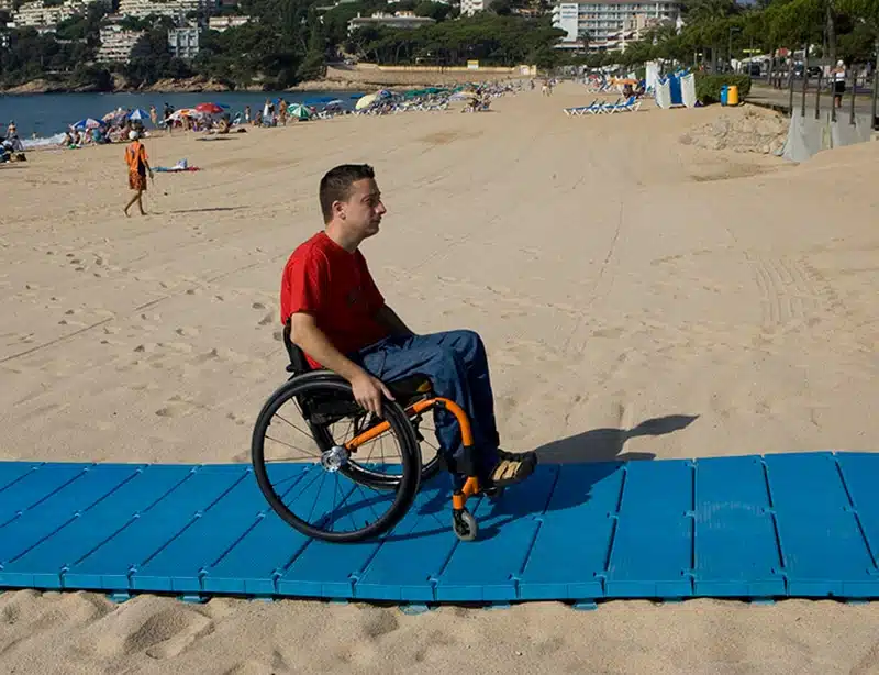 La imagen muestra a una persona utilizando una silla de ruedas sobre una pasarela de playa azul colocada sobre la arena de una playa. La pasarela parece proporcionar un camino estable para la silla de ruedas sobre la arena, facilitando el acceso al área de la playa. En el fondo, hay personas realizando diversas actividades como caminar y tomar el sol.