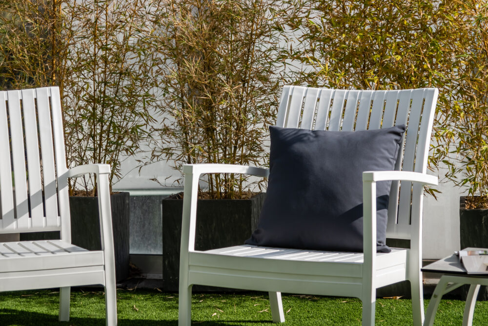 Se puede observar en la imagen dos sillones de color blanco, de diseño moderno y elegante con un cojin negro encima del sillón, de fondo se ve vegetación